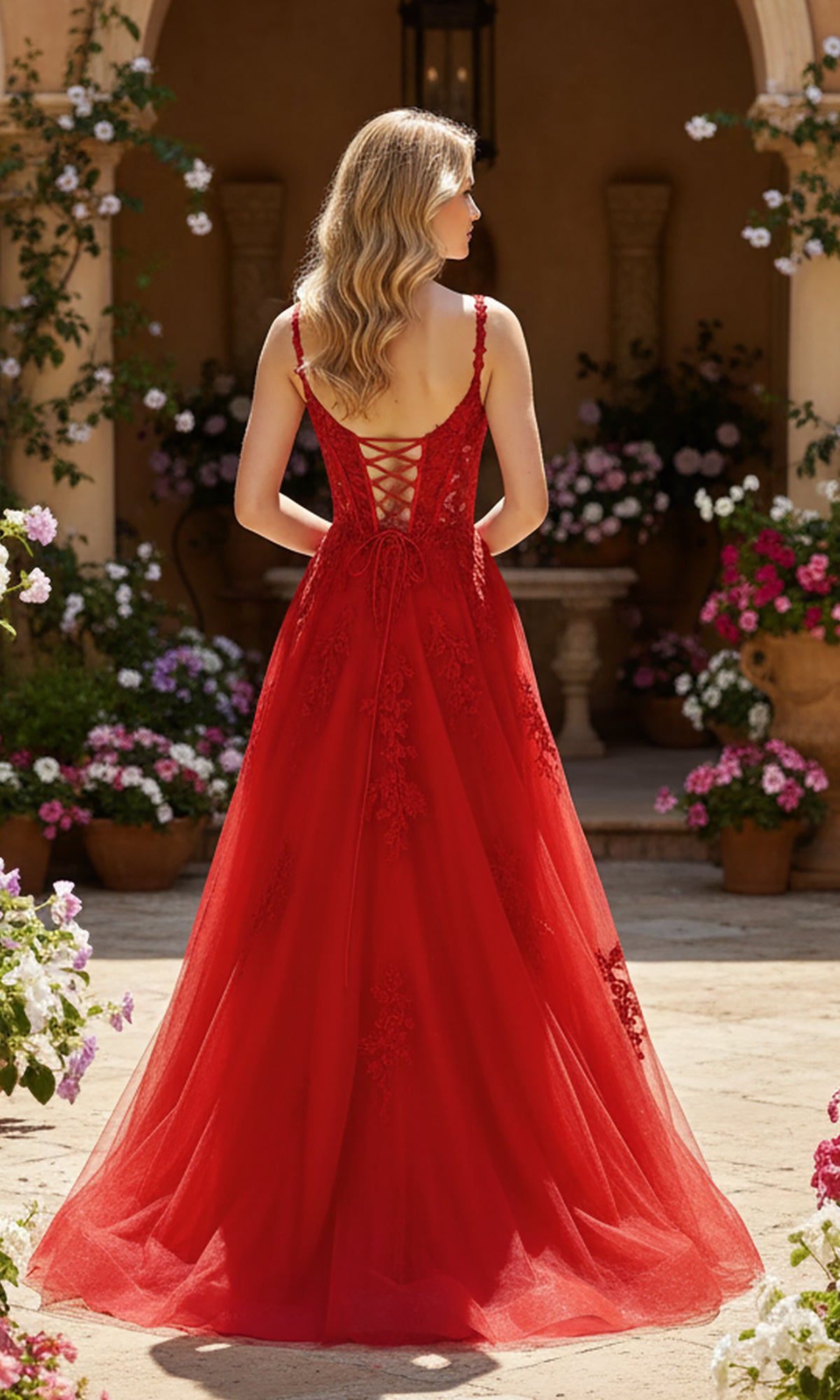 A woman with wavy blonde hair wears the Long Prom Dress: PromGirl Exclusive 250261, a red lace applique gown with a lace-up back, standing outdoors among potted flowers and plants.